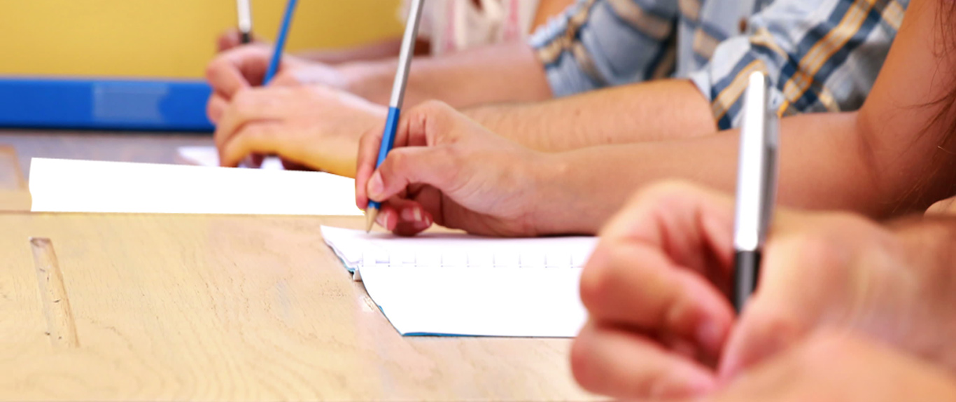 Close-up of several hands writing with pens and pencils on paper at a wooden desk, warm yellow tones, focused studious mood